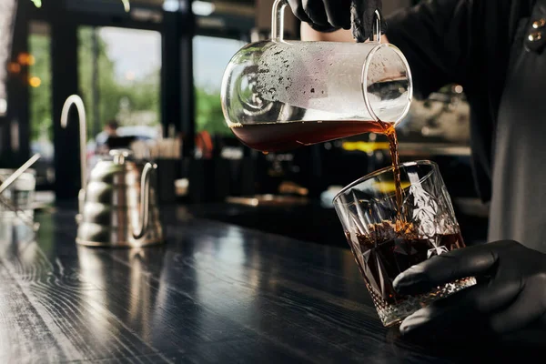 Cropped view of barista holding coffee pot and pouring freshly brewed espresso into crystal glass — Stock Photo