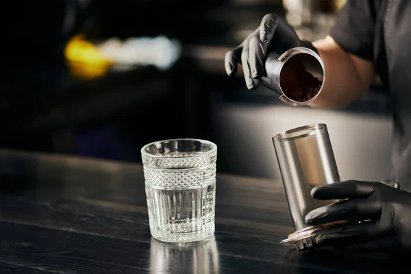Vista parcial de barista en vidrio de látex negro sosteniendo prensa aerodinámica con café cerca de vidrio sobre mesa negra - foto de stock