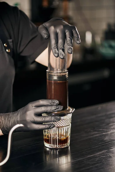 Cropped view of barista in black gloves pressing ground coffee in aero press while preparing espresso — Stock Photo