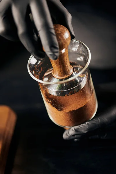 Vista ritagliata di barista in guanti di lattice nero premendo caffè macinato in gocciolamento freddo caffettiera in caffè — Foto stock