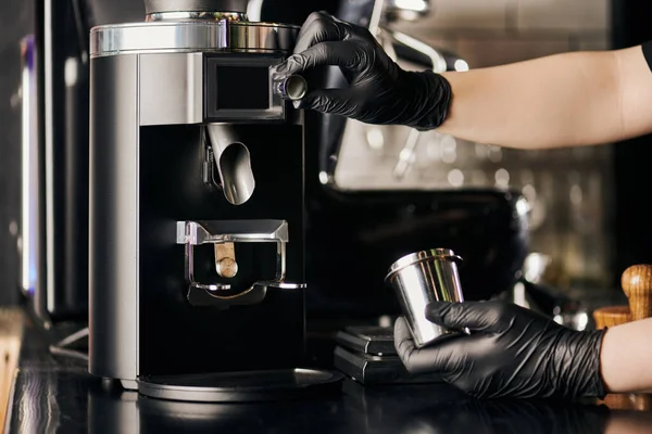 Barista em luvas de látex preto operando moedor de café elétrico e segurando copo de medição metálica — Fotografia de Stock