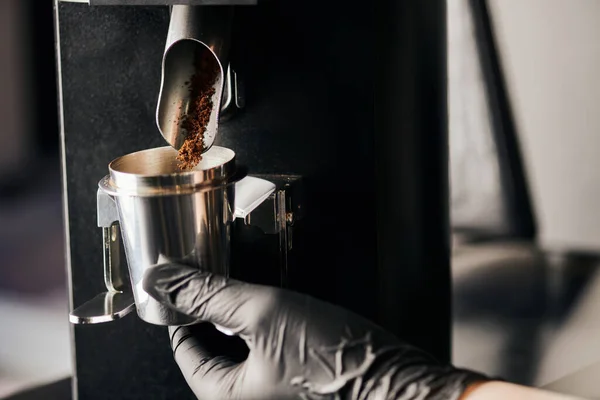 Barista in schwarzem Latex-Handschuh mit Messbecher in der Nähe von gemahlenem Kaffee und elektrischer Kaffeemühle — Stockfoto