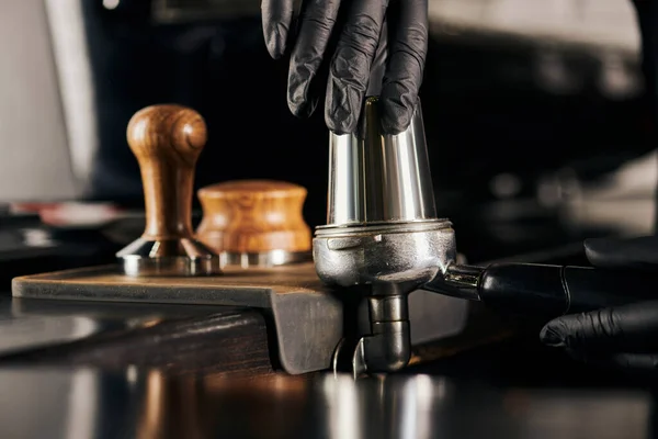 Cropped view of barista in black latex glove pressing coffee in portafiler while preparing espresso — Stock Photo