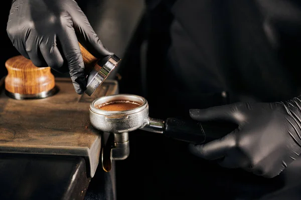 Cropped view of barista in black latex gloves pressing ground coffee in portafilter, coffee shop — Stock Photo