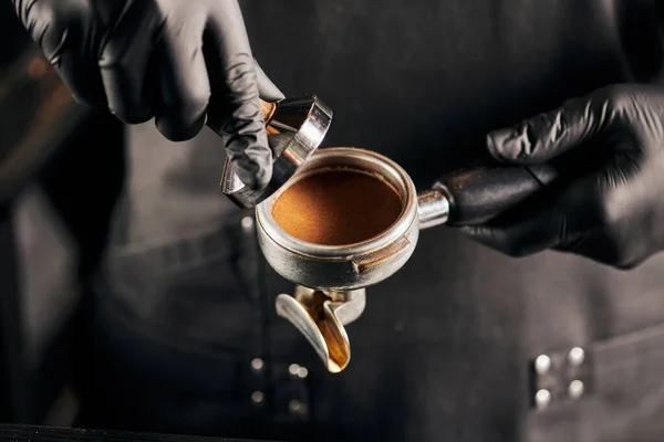 Cropped view of tamper with ground coffee and portafilter in hands of barista in black gloves — Stock Photo