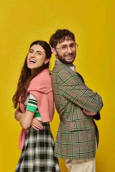 Счастливые стильные мужчина и женщина, стоя спиной к спине, позируя, желтый фон, студенческий наряд, смех — стоковое фото