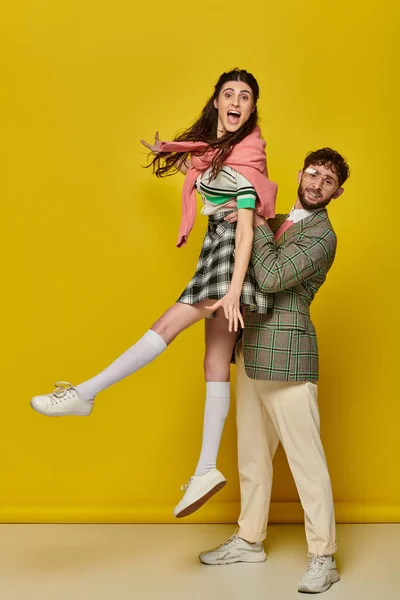 Coppia allegra divertirsi, uomo in occhiali sollevamento eccitato giovane donna su sfondo giallo, studenti — Foto stock