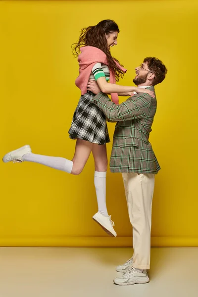 Alegre pareja divirtiéndose, hombre en gafas levantando excitado joven mujer en amarillo telón de fondo, estudiantes - foto de stock