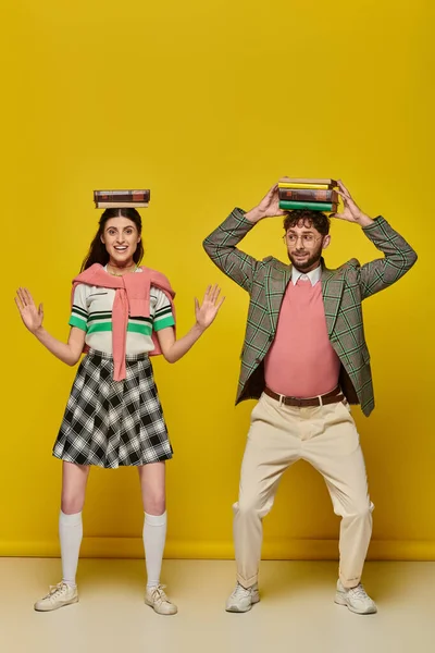 Funny couple, happy young man and woman standing with books on heads on yellow backdrop, students — Stock Photo