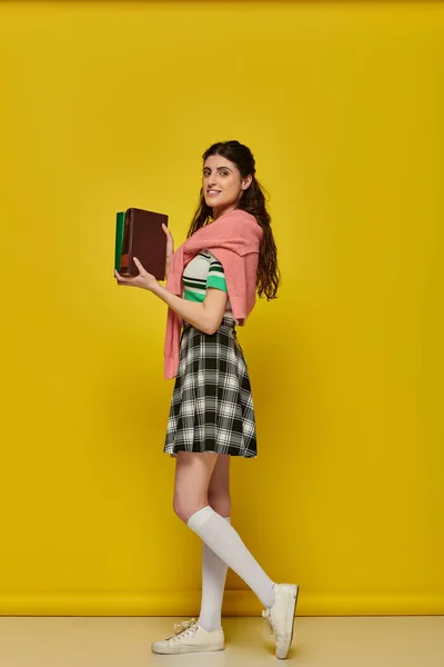 Étudiant joyeux debout avec des livres sur fond jaune, jeune femme en jupe, tenue universitaire — Stock Photo