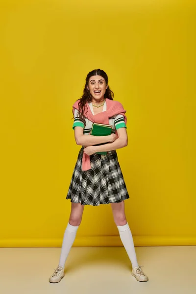 Excited young woman in skirt standing with books on yellow backdrop, happy student, college outfit — Stock Photo
