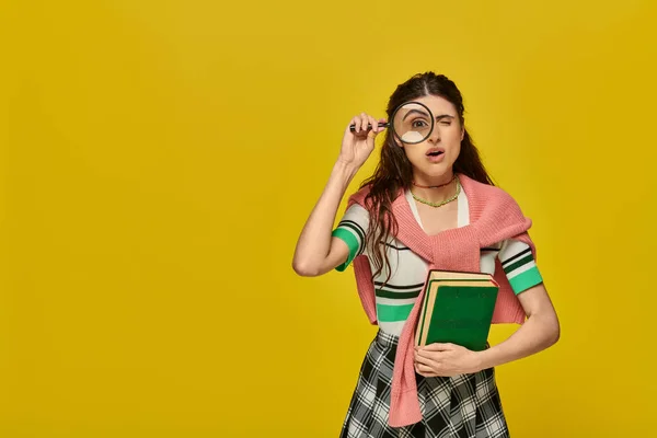 Estudiante curioso sosteniendo libros y lupa, zoom, descubrimiento, mujer joven en traje de la universidad, amarillo - foto de stock