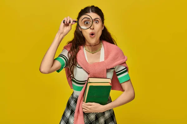 Curiosa joven sosteniendo libros y lupa, zoom, descubrimiento, estudiante en traje universitario, amarillo - foto de stock
