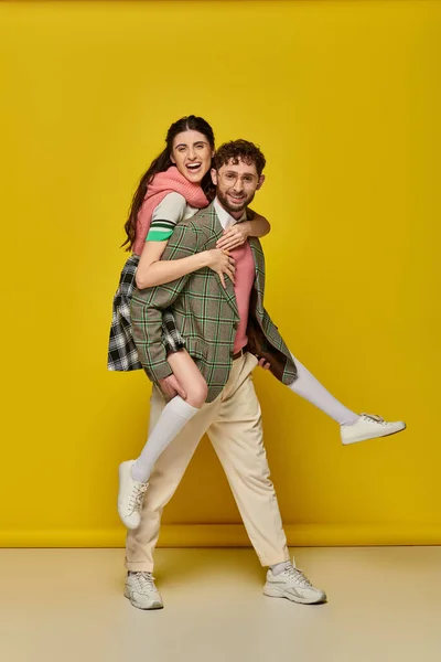 Aufgeregtes Paar, bärtiger junger Mann huckepack brünette Frau auf gelbem Hintergrund, lustige Studenten — Stockfoto