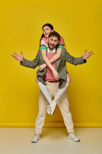 Excited couple, funny, young man piggybacking brunette woman on yellow backdrop, student outfits — Stock Photo