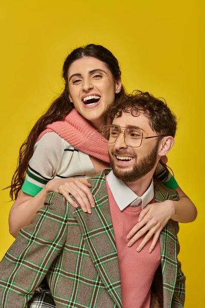 Divertidos estudiantes, hombre alegre piggybacking mujer joven en amarillo telón de fondo, trajes universitarios, pareja - foto de stock
