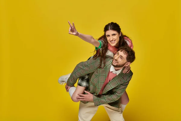 Estudantes engraçados, homem alegre piggybacking mulher no fundo amarelo, v sinal, roupas da faculdade, casal — Fotografia de Stock