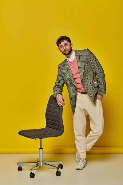 Бородатий чоловік стоїть з рукою в кишені біля офісного крісла, жовтий фон, студент в окулярах — стокове фото