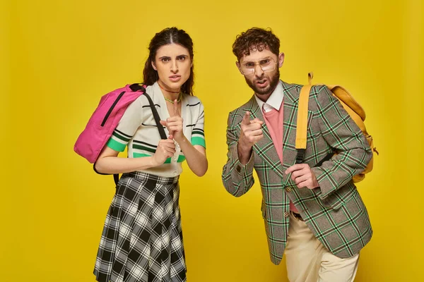 Angry students standing with backpacks, looking at camera, pointing with finger, yellow backdrop — Stock Photo