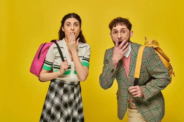 Shocked students standing with backpacks, looking at camera, covering mouth, yellow backdrop, wow — Stock Photo