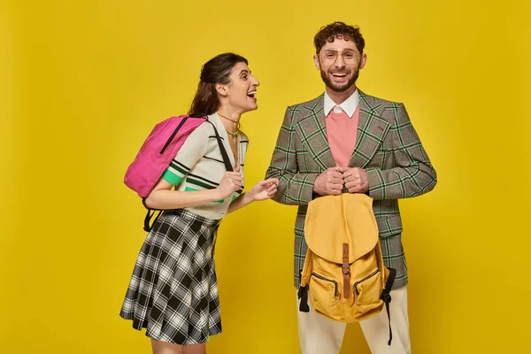 Estudiantes divertidos de pie con mochilas, mirando a la cámara, sonriendo, fondo amarillo, ropa académica - foto de stock