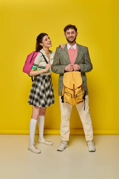 Estudiantes de pie con mochilas, mirando a la cámara, sonriendo, fondo amarillo, ropa académica, estilo - foto de stock