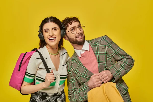 Femme gaie dans des écouteurs sans fil debout près de l'homme barbu, les étudiants tenant des sacs à dos, la musique — Photo de stock