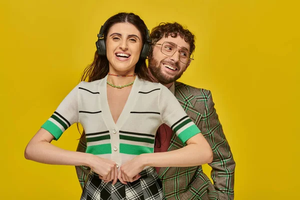 Colegio, mujer feliz en auriculares escuchando música cerca de hombre barbudo, estudiantes divertidos, fondo amarillo - foto de stock