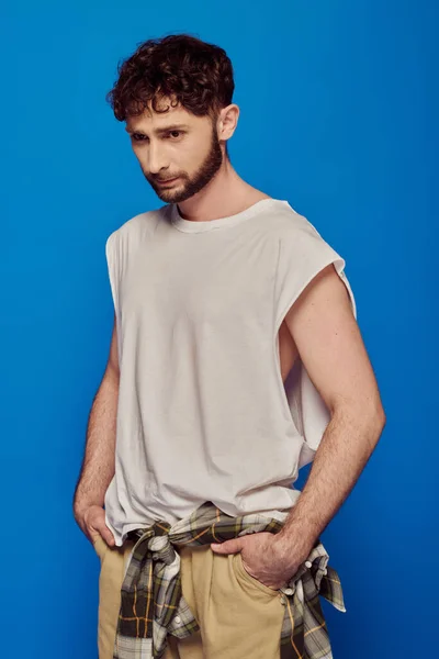 Bearded and curly man standing with hands in pockets on blue background, tank top, male fashion — Stock Photo