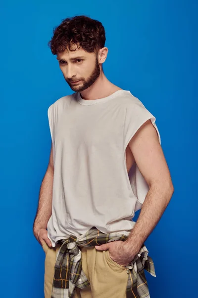 Hombre barbudo y rizado de pie con las manos en los bolsillos sobre fondo azul, camiseta sin mangas, moda masculina - foto de stock