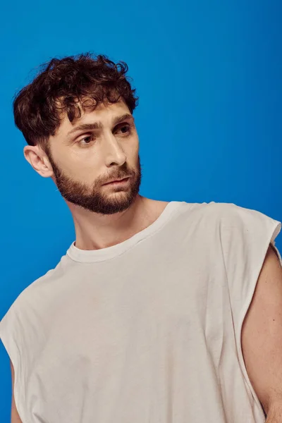 Bearded and curly man posing on blue background, white tank top, male fashion, looking away — Stock Photo