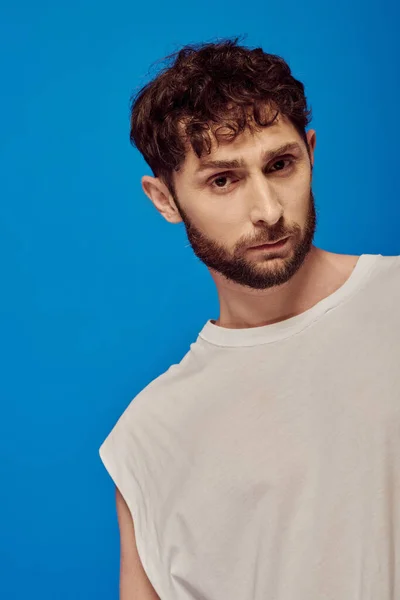 Bearded and curly man posing on blue background, white tank top, male fashion, looking at camera — Stock Photo