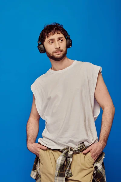 Bearded man in wireless headphones listening music on blue background, white tank top, male fashion — Stock Photo
