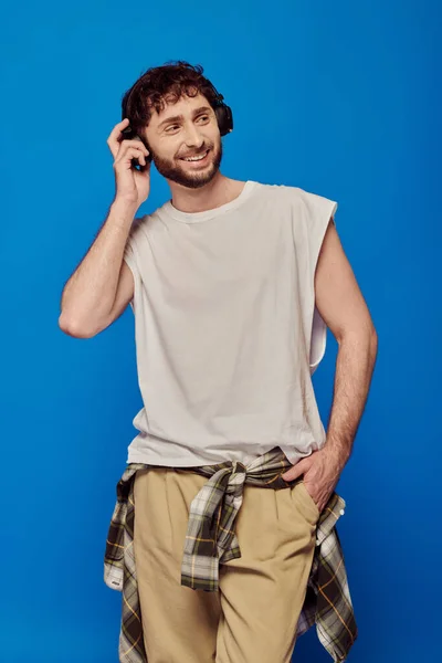 Uomo allegro in cuffie wireless ascoltare musica su sfondo blu, mano nella tasca, moda maschile — Foto stock