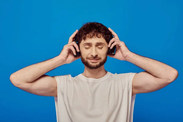 Pleased man in wireless headphones listening music on blue background, white tank top, closed eyes — Stock Photo