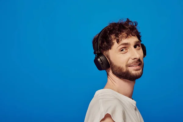 Hombre positivo en auriculares inalámbricos escuchando música sobre fondo azul, moda masculina, audio - foto de stock