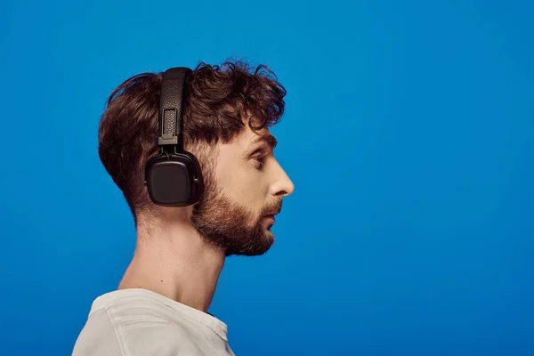 Seitenansicht, bärtiger Mann mit drahtlosen Kopfhörern, der Musik auf blauem Hintergrund hört, Männermode — Stockfoto
