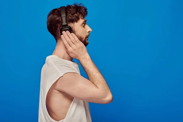 Side view, bearded man in wireless headphones listening music on blue background, summer fashion — Stock Photo