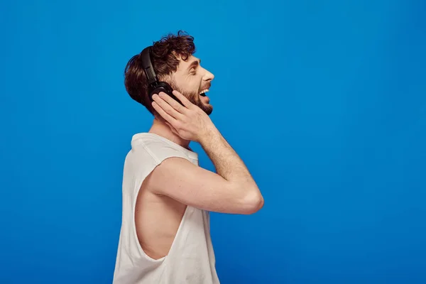Vista lateral, homem alegre em fones de ouvido sem fio ouvir música no fundo azul, moda verão — Fotografia de Stock