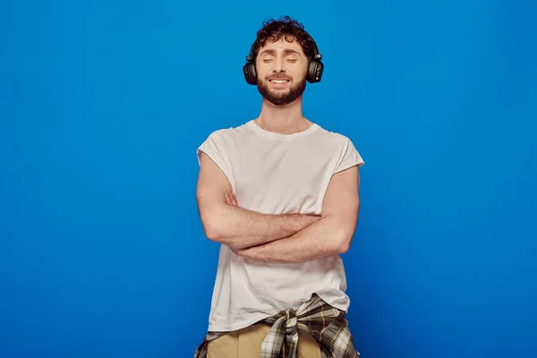 Hombre positivo en auriculares inalámbricos escuchando música sobre fondo azul, ojos cerrados, moda de verano - foto de stock