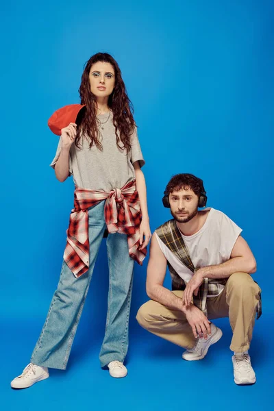 Couple branché, écouteurs, musique, street wear, maquillage audacieux, femme avec casquette de baseball, fond bleu — Photo de stock