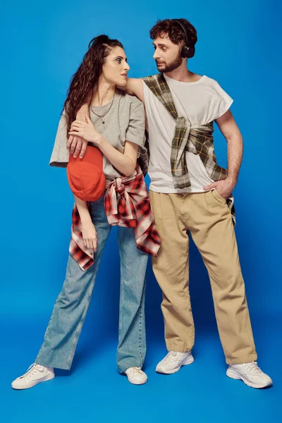 College couple, headphones, music, street wear, bold makeup, woman with baseball cap, blue backdrop — Stock Photo