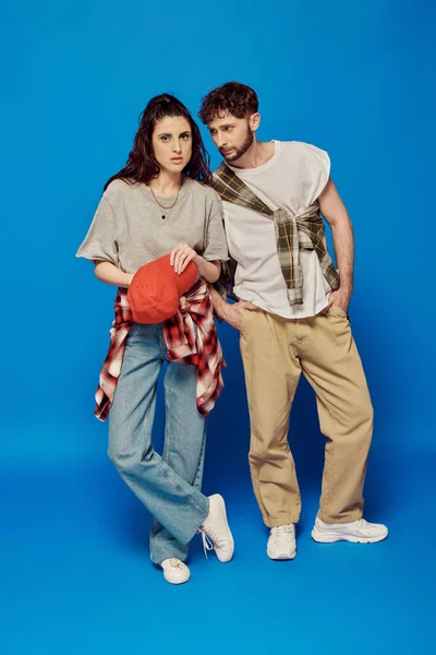 Пара коледжу позує у вуличному одязі на синьому фоні, жінка зі сміливим макіяжем, бейсбольна шапка — стокове фото