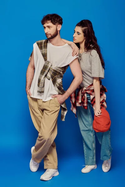 Couple posant en tenue de rue sur fond bleu, femme au maquillage audacieux, homme barbu, casquette de baseball — Photo de stock