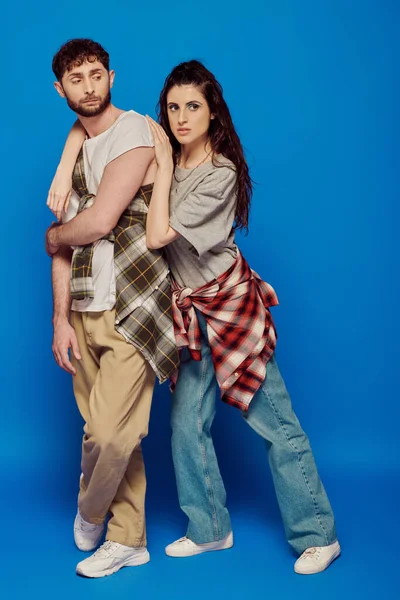 Couple posing in street wear, blue backdrop, woman with bold makeup leaning on bearded man, trend — Stock Photo