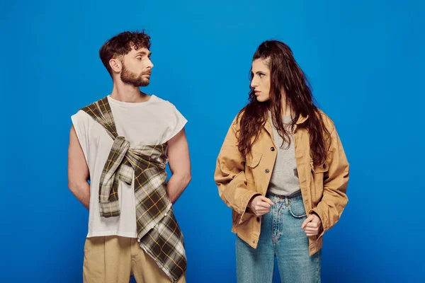 Stylish couple in casual wear looking at each other, blue backdrop, woman standing with bearded man — Stock Photo