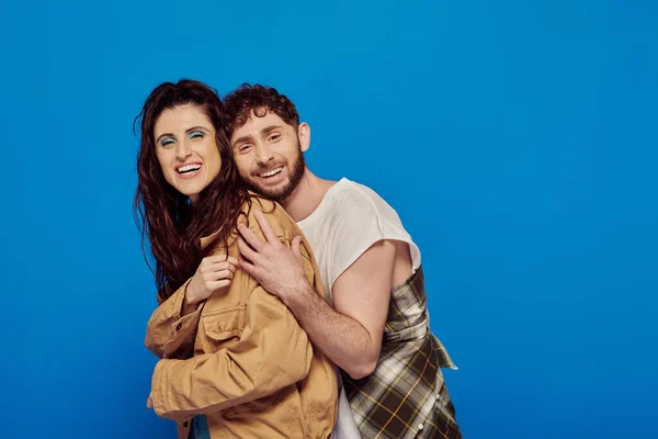 Cheerful couple in casual wear posing on blue backdrop, bearded man hugging woman, bold makeup — Stock Photo