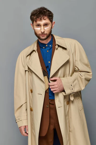 Vêtements de dessus, homme à la mode en lunettes et trench coat posant sur fond gris, couches confortables, mode automne — Photo de stock