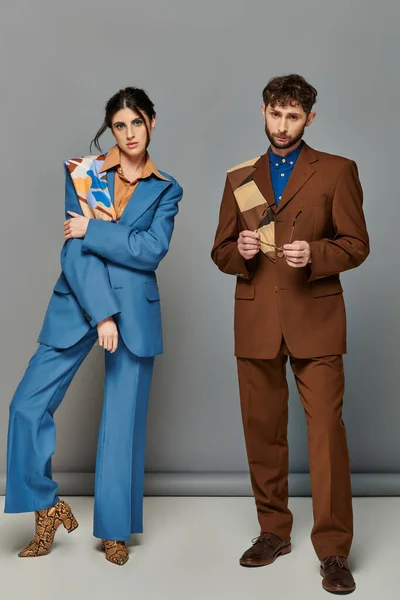 Мужчина и женщина в костюмах, стоящие на сером фоне, модные съемки, пара, посмотрите в камеру — стоковое фото