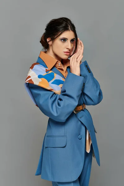 Portrait, femme brune au maquillage audacieux, rêveuse, mannequin en costume bleu, tenue formelle, fond gris — Photo de stock